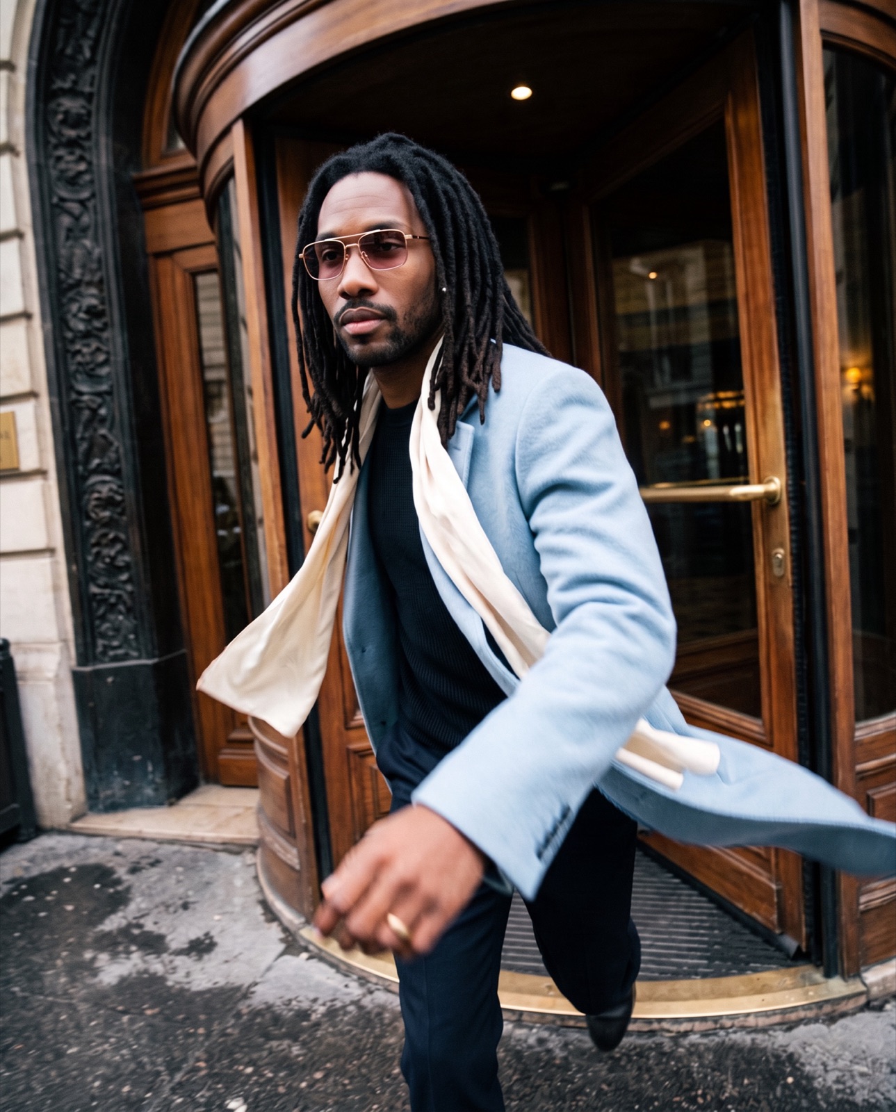 Man with long dreadlocks wearing rose-tinted aviator glasses stepping through a hotel revolving door
