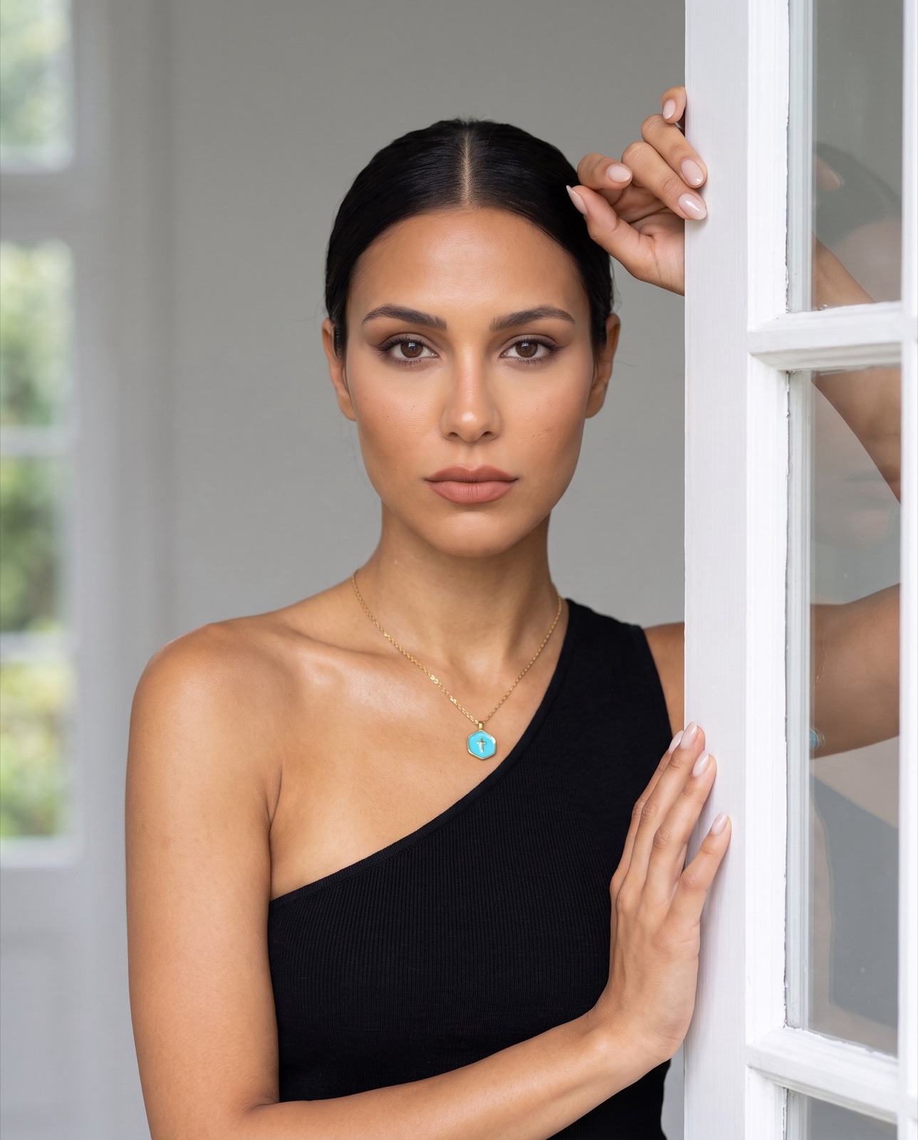 Model wearing a gold pendant necklace and a black one-shoulder top by a window